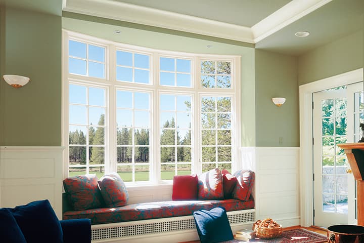 Bright modern living room with large multi-pane windows overlooking forest, featuring red window seat cushions and sage green walls